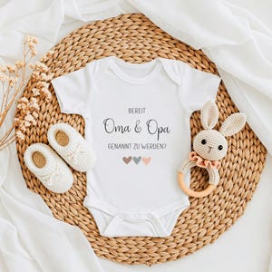 K&ouml;nnte beinhalten: Ein wei&szlig;er kurz&auml;rmeliger Babybody mit dem schwarzen Text "BEREIT Oma & Opa GENANNT ZU WERDEN?" und drei kleinen Herzen in gedeckten Farben. Der Body wird von einem Paar wei&szlig;er Stricksch&uuml;hchen und einer geh&auml;kelten Hasenrassel begleitet.