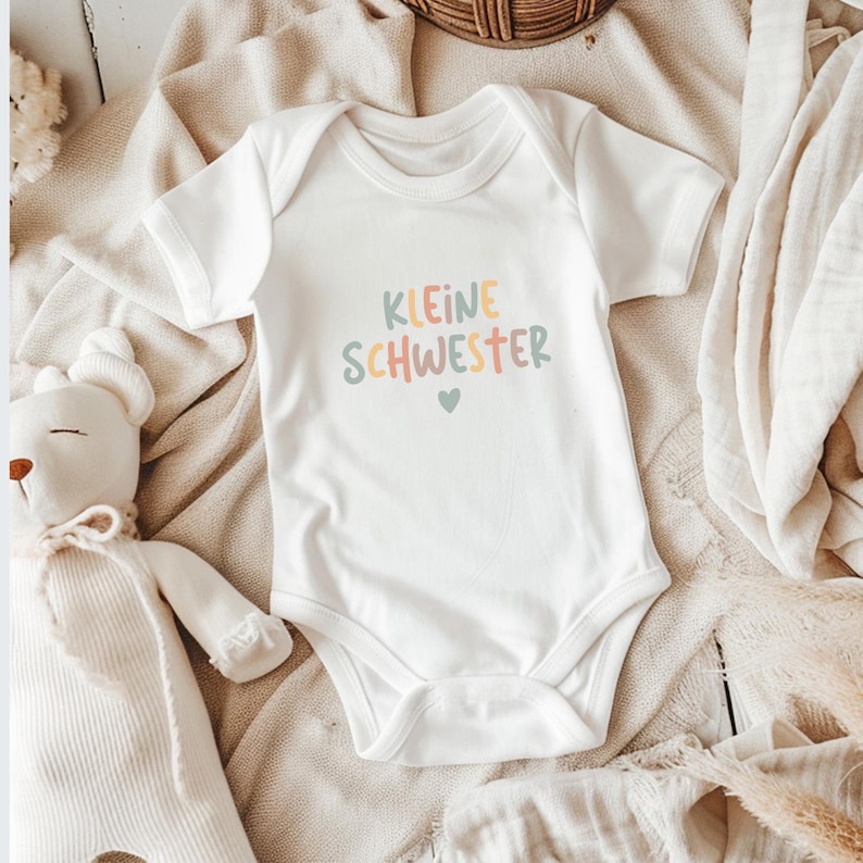 May include: A white short-sleeved baby bodysuit featuring the text "KLEINE SCHWESTER" printed in multicolored pastel letters, with a small heart icon below the text. The garment has a round neckline and standard leg openings.