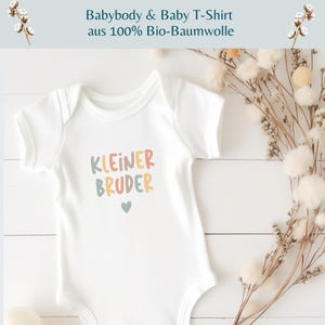 May include: A white short-sleeved baby bodysuit featuring the text "KLEINER BRUDER" in multicolored letters and a small heart icon below the text. The garment is displayed alongside dried floral stems on a white wooden surface.