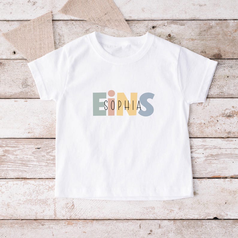 May include: A white short-sleeved t-shirt featuring the word EINS in large, muted pastel block letters in sage green, peach, yellow, and dusty blue, with the name SOPHIA printed in a thin, black, sans-serif font centered across the letters.