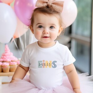 May include: A white short-sleeved bodysuit featuring the word EINS in muted pastel block letters with the name ANNABEL printed in black across the center. The garment is paired with a light pink tulle skirt and a glittery rose gold hair bow.