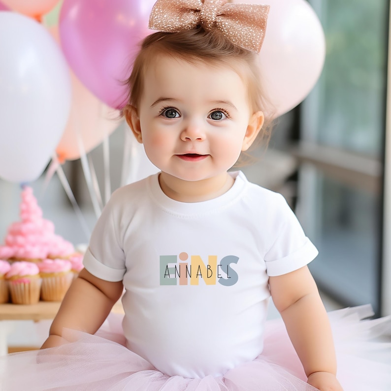 May include: A white short-sleeved bodysuit featuring the word EINS in muted pastel block letters with the name ANNABEL printed in black across the center. The garment is paired with a light pink tulle skirt and a glittery rose gold hair bow.