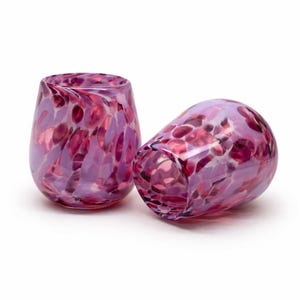 May include: Two stemless wine glasses with a speckled design. The glasses are a mix of purple and pink hues, with a rounded shape. The glass on the left is upright, while the other is tilted on its side.