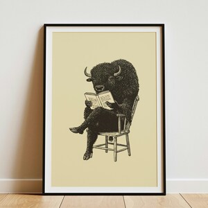Könnte beinhalten: Ein gerahmter Kunstdruck mit einer schwarzen Tuschezeichnung eines Bisons, das auf einem Holzstuhl sitzt und ein offenes Buch liest. Das Bison ist mit übereinandergeschlagenen Beinen dargestellt und hält das Buch mit seinen Vorderhufen vor einem beigen Hintergrund.