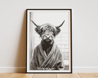 May include: Black and white framed print of a Highland cow wearing a bathrobe. The cow has large horns and is centered in the frame. The background shows a tiled wall. The artwork is displayed on a white wall.