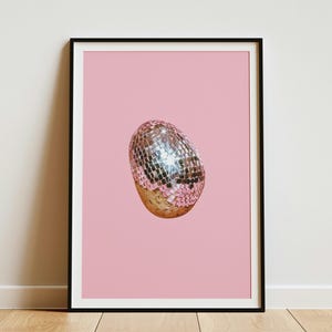 Op de afbeelding: Een ingelijste kunstdruk met een gecentreerd, eivormig object dat aan de bovenkant is bedekt met reflecterende zilveren spiegeltjes en aan de onderkant een gladde, lichtbruine textuur heeft, tegen een effen, lichtroze achtergrond.