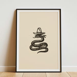 May include: A framed wall art print featuring a black ink illustration of a coiled snake wearing a cowboy hat on a solid light beige background. The artwork is enclosed in a thin black frame, presenting a centred graphic design.