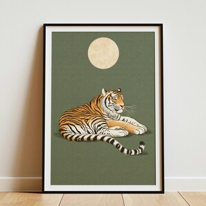 May include: A framed wall art print featuring an illustration of an orange and black striped tiger resting on a solid olive green background. Above the tiger, a large, pale yellow circular moon is centred in the upper portion of the composition.