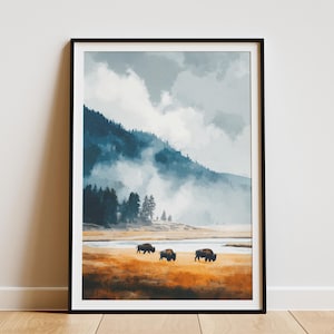 May include: Framed art print featuring a landscape scene with bison. The artwork depicts a mountain range with a misty atmosphere, a river, and three bison grazing in a field of golden grass. The print is in a black frame.