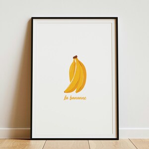 Könnte beinhalten: Ein gerahmter Kunstdruck mit einer gelben Bananenillustration, die mittig auf einem weißen Hintergrund platziert ist. Unter der Banane steht der Text la bananne in einer gelben Schreibschrift. Das Bild ist in einem dünnen schwarzen Rahmen.