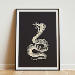 May include: A framed wall art print featuring a high-contrast illustration of a coiled cobra snake with its mouth open, displaying fangs. The snake is rendered in cream-colored lines against a solid black background, enclosed in a thin black frame.