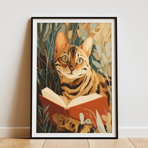 May include: A framed art print featuring a Bengal cat reading a book. The cat has a brown and black striped pattern, with large green eyes. The book is open, with white pages and a red cover. The background includes green and beige foliage.