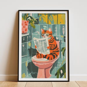 May include: A framed art print depicts a whimsical scene of a cat reading a newspaper while sitting on a toilet. The cat has orange and black stripes. The bathroom has teal tiles and a pink toilet.