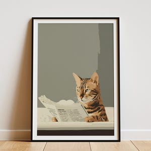 May include: Framed art print featuring a Bengal cat reading a newspaper. The cat has brown and black markings and is set against a muted gray background. The print is in a black frame and is displayed on a white wall.
