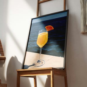 May include: A framed wall art print depicting a tall glass filled with a bright orange liquid and garnished with an orange slice. The glass sits on a light-colored surface against a dark blue sea background, casting a distinct shadow.