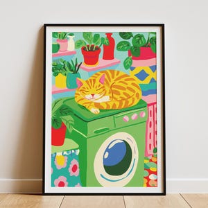 May include: A framed art print shows a sleeping ginger cat on a green washing machine. The background features shelves with potted plants and colourful decorative elements, creating a cosy, whimsical scene. The artwork is framed in a black border.