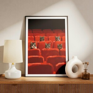 May include: A framed wall art print featuring a digital illustration of five cats sitting in red theater seats. The cats are positioned in different rows, looking forward against a dark background. The print is displayed in a thin black frame.