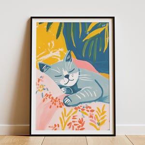 Op de afbeelding: Een ingelijste kunstprint met een illustratie van een slapende kat. De kat is lichtblauw en rust op een roze kussen met bloemmotieven. De achtergrond bevat gele, blauwe en groene penseelstreken, wat een levendige scène creëert.