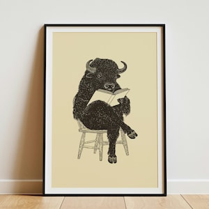 May include: A framed wall art print featuring a black ink illustration of a bison sitting on a wooden chair while reading an open book. The bison is depicted with detailed fur texture, and the background of the print is a solid light beige color.