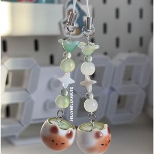 Könnte beinhalten: Zwei Perlen-Telefonanhänger mit weißen Kordeln. Jeder Anhänger hat grüne und weiße Perlen mit Stern- und Blumenakzenten. Der Boden jedes Anhängers hat eine kleine, runde, weiß-orangefarbene Keramikperle. Der Text auf dem Bild lautet "JELLYBELLYJULEZ".