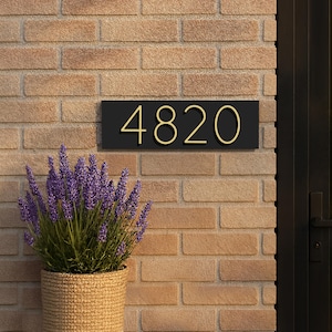 May include: A black rectangular address plaque with the number 4820 in gold lettering is mounted on a brick wall. A woven basket with purple lavender plants sits below the plaque, near a black door.
