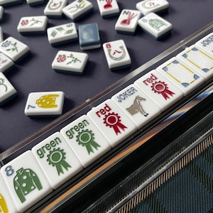 May include: A close-up of a Mahjong set with white tiles featuring various symbols and characters. The tiles are arranged on a dark blue surface, with some tiles in a clear plastic holder. The set includes tiles marked with "green", "red", and "Joker".
