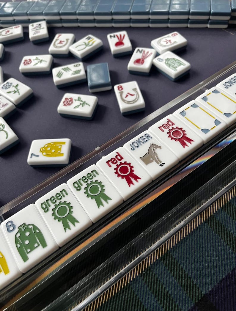 May include: A close-up of a Mahjong set with white tiles featuring various symbols and characters. The tiles are arranged on a dark blue surface, with some tiles in a clear plastic holder. The set includes tiles marked with "green", "red", and "Joker".