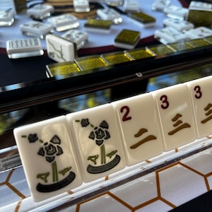 May include: Close-up of a Mahjong set with white tiles featuring floral designs and numbers. The tiles are arranged on a black surface with a gold-colored border. Other tiles are scattered in the background.