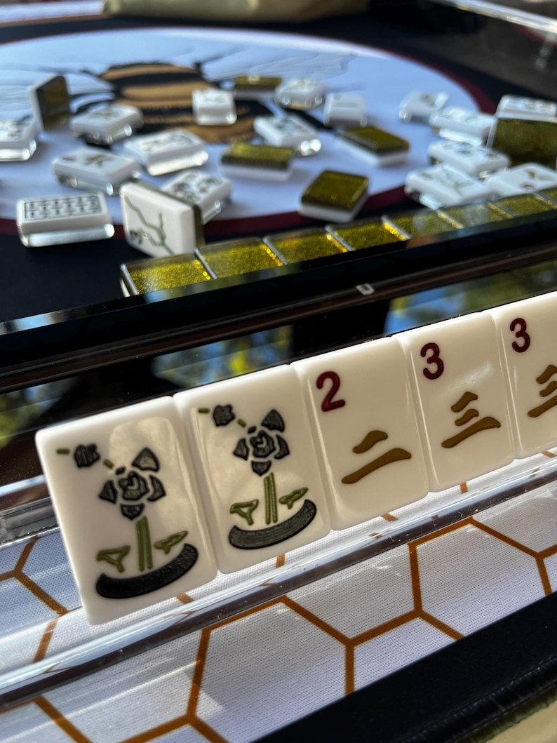 May include: Close-up of a Mahjong set with white tiles featuring floral designs and numbers. The tiles are arranged on a black surface with a gold-colored border. Other tiles are scattered in the background.