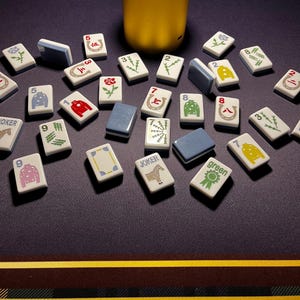 May include: A collection of Mahjong tiles with various symbols and numbers, including Jokers and green tiles. The tiles are white with blue edges and are scattered on a dark surface. A gold-colored cylindrical object is in the background.