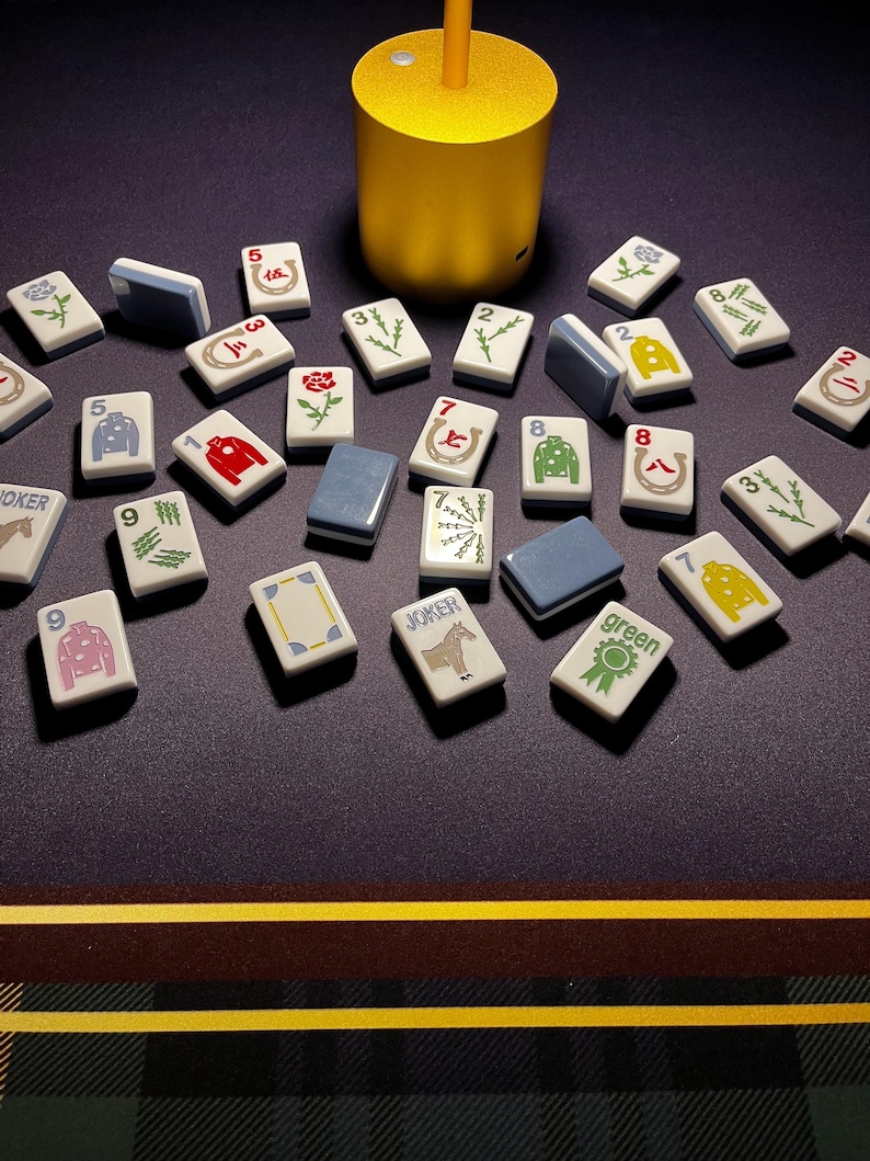 May include: A collection of Mahjong tiles with various symbols and numbers, including Jokers and green tiles. The tiles are white with blue edges and are scattered on a dark surface. A gold-colored cylindrical object is in the background.