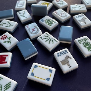 May include: A collection of Mahjong tiles scattered on a dark blue surface. The tiles are white with various symbols and numbers, including roses, horseshoes, and the word "Joker." Some tiles have a solid blue back.