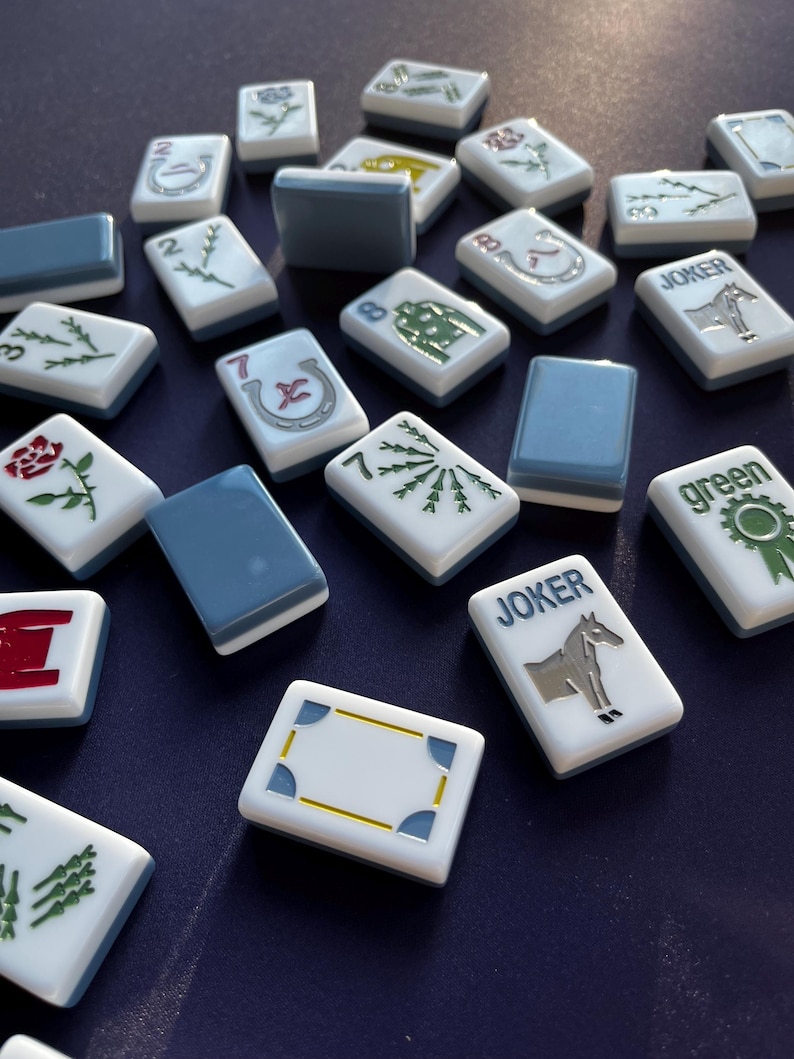 May include: A collection of Mahjong tiles scattered on a dark blue surface. The tiles are white with various symbols and numbers, including roses, horseshoes, and the word "Joker." Some tiles have a solid blue back.