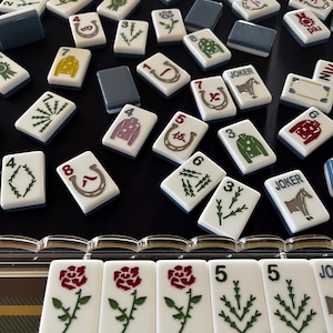 May include: A collection of white Mahjong tiles with various symbols and numbers, including roses, horseshoes, and jackets. The tiles have a blue border and are scattered on a black surface. Some tiles have the word "JOKER" on them.