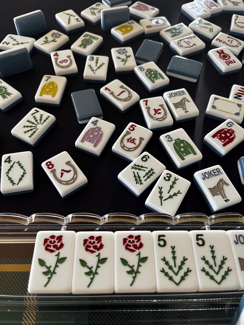 May include: A collection of white Mahjong tiles with various symbols and numbers, including roses, horseshoes, and jackets. The tiles have a blue border and are scattered on a black surface. Some tiles have the word "JOKER" on them.