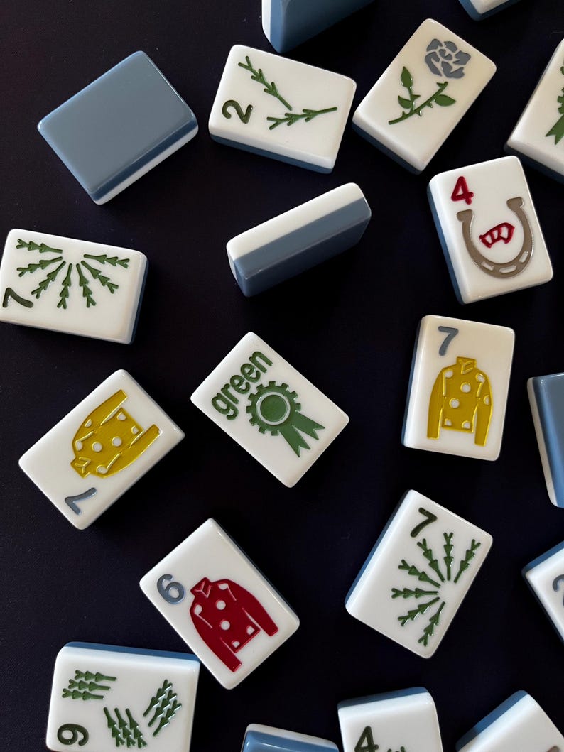 May include: A collection of white and blue Mahjong tiles with various symbols and numbers. The tiles feature green, red, and yellow designs, including a horseshoe, a rose, and a ribbon. The word "green" is printed on one tile.