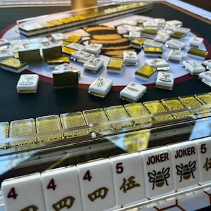 May include: A Mahjong set with white tiles, gold accents, and a black and gold playing surface. The tiles display numbers, symbols, and the word "Joker". The set includes racks and a golden cylindrical object.