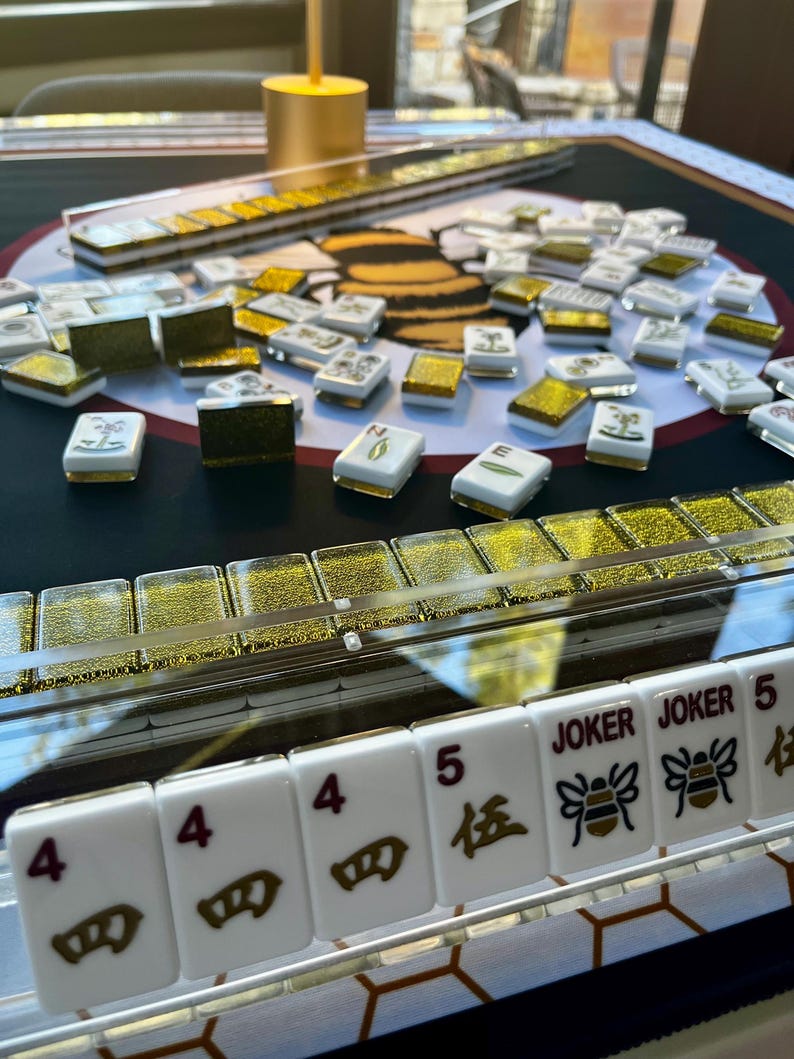 May include: A Mahjong set with white tiles, gold accents, and a black and gold playing surface. The tiles display numbers, symbols, and the word "Joker". The set includes racks and a golden cylindrical object.
