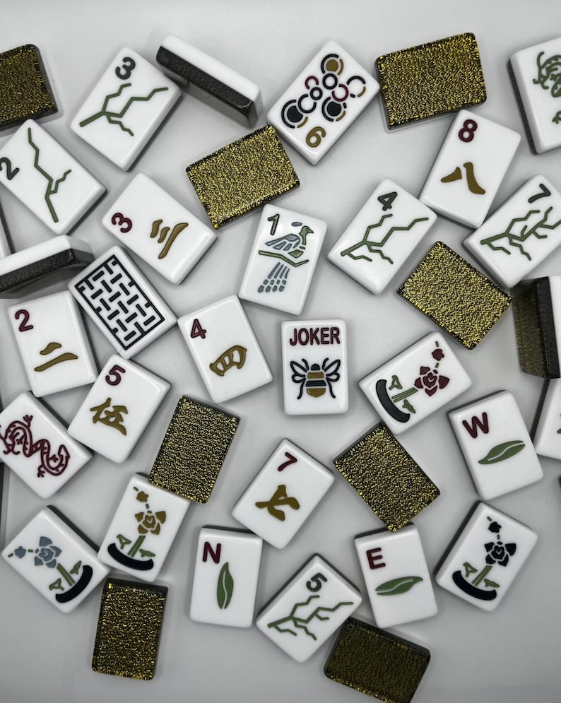 May include: A collection of white Mahjong tiles with black, green, and brown symbols and numbers. Some tiles feature floral designs, while others display the letters "N", "E", and "W". Several gold-colored, textured tiles are also present. One tile reads "JOKER".