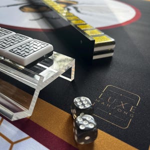 May include: A close-up of a Mahjong set with black dice, white tiles, and a black and gold playing surface. The set includes a clear acrylic tile holder and a black and gold tile rack. The words "LUXE MAHJONG" are printed on the surface.