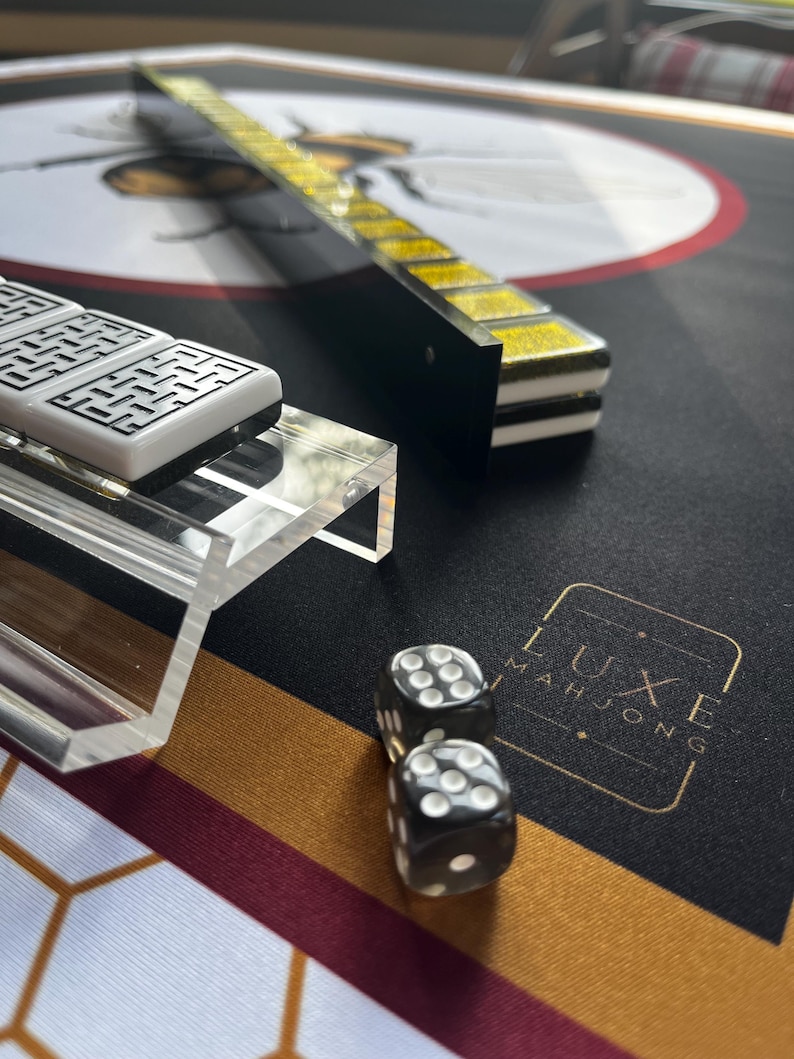 May include: A close-up of a Mahjong set with black dice, white tiles, and a black and gold playing surface. The set includes a clear acrylic tile holder and a black and gold tile rack. The words "LUXE MAHJONG" are printed on the surface.