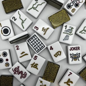 May include: A collection of white Mahjong tiles with various symbols and numbers in black, green, and brown. Some tiles feature Chinese characters and a "JOKER" tile with a bee illustration. Several gold-colored tiles are also present.