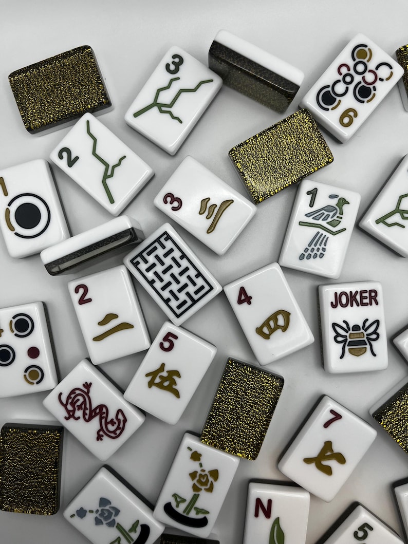 May include: A collection of white Mahjong tiles with various symbols and numbers in black, green, and brown. Some tiles feature Chinese characters and a "JOKER" tile with a bee illustration. Several gold-colored tiles are also present.