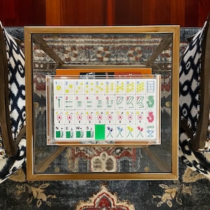 May include: A clear acrylic case containing a Mahjong set with white tiles. The tiles display various symbols, numbers, and characters in green, red, and black. The set is on a glass-topped table with a gold-colored frame.