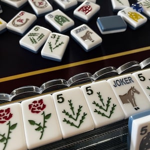 May include: A collection of white Mahjong tiles with various symbols and numbers, including roses, horses, and green plant designs. Some tiles have the word "JOKER" on them. The tiles are arranged on a dark surface.
