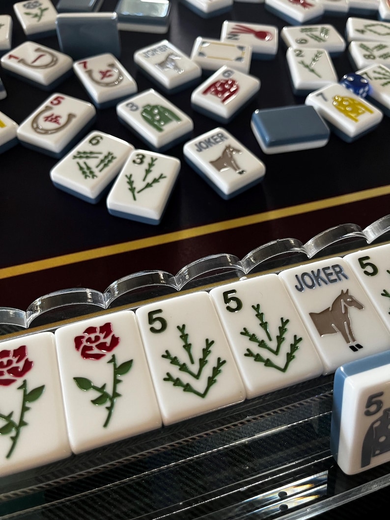 May include: A collection of white Mahjong tiles with various symbols and numbers, including roses, horses, and green plant designs. Some tiles have the word "JOKER" on them. The tiles are arranged on a dark surface.