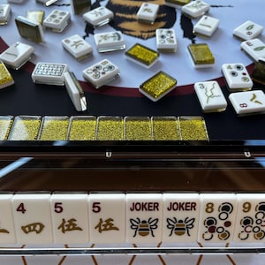 May include: A close-up of a Mahjong set with white tiles featuring black and gold symbols and numbers. The set includes "JOKER" tiles with bee graphics. A large bee illustration is in the background.