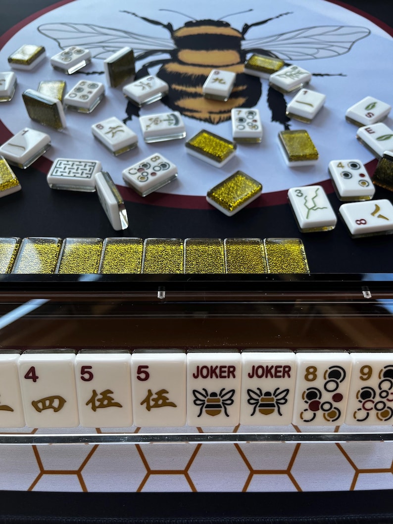 May include: A close-up of a Mahjong set with white tiles featuring black and gold symbols and numbers. The set includes "JOKER" tiles with bee graphics. A large bee illustration is in the background.