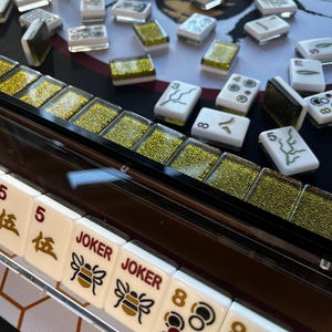 May include: A close-up of a Mahjong set with white tiles featuring black and gold markings. Some tiles have the word "JOKER" and a bee graphic. The set includes a clear acrylic tile holder and is set on a black surface with a bee design.
