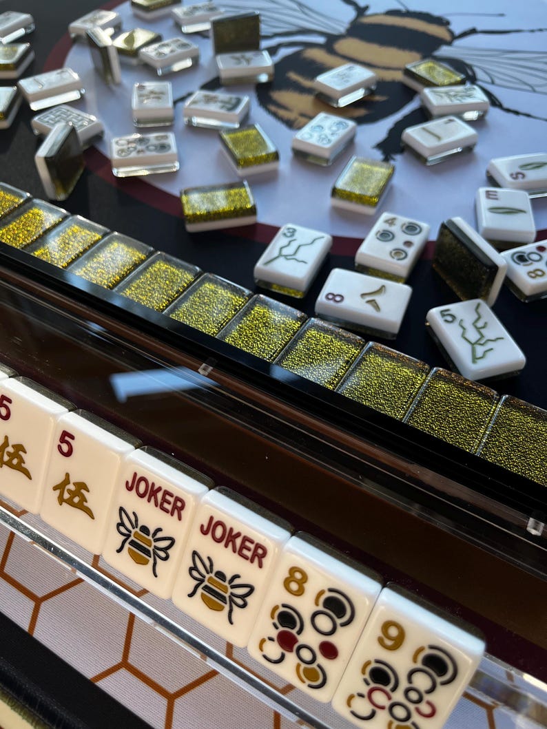 May include: A close-up of a Mahjong set with white tiles featuring black and gold markings. Some tiles have the word "JOKER" and a bee graphic. The set includes a clear acrylic tile holder and is set on a black surface with a bee design.